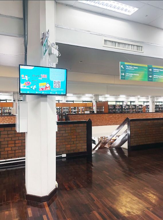Students pass a wall mounted digital signage screen near the library circulation area, where announcements and course information rotate throughout the day.